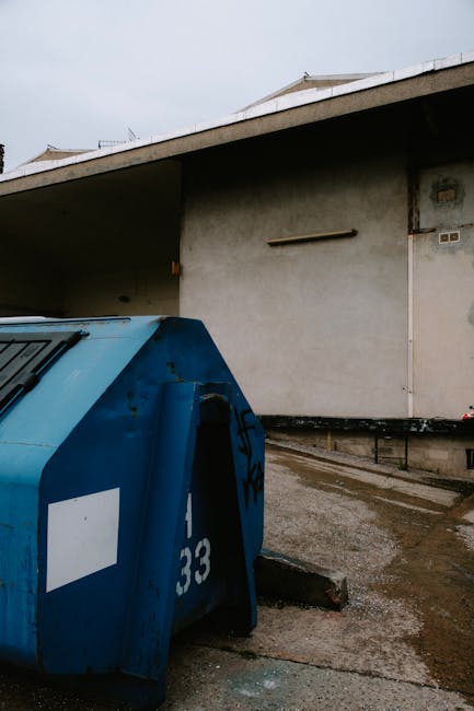 In the foreground, a large blue skips positioned on a paved surface, showing signs of rust and graffiti markings; its curved metal panels and sliding doors are visibly weathered and slightly dented. B